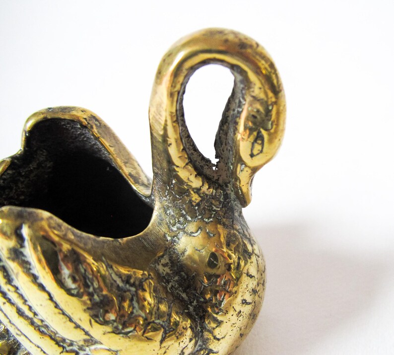 May include: A vintage brass swan figurine with a textured surface. The swan has a curved neck and a hollowed-out body, possibly for holding small items. The brass has a warm, golden hue, and the swan is set against a white background.