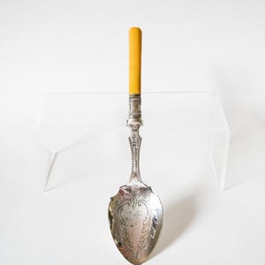 May include: A silver spoon with an ornate design on the bowl and handle. The handle has a yellow grip. The spoon is likely vintage or antique, and is displayed on a clear surface against a white background.