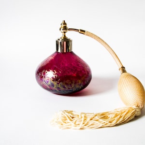 May include: A bulbous magenta glass perfume bottle with a gold-toned metal nozzle and a beige fabric-covered atomiser bulb. The glass features a swirling iridescent pattern at the base, and the atomiser includes a long, matching beige tassel.
