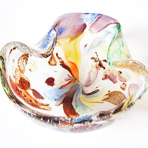 May include: A decorative glass bowl with a unique, freeform shape. The bowl is clear with colorful swirls of orange, yellow, blue, and purple. The glass has a speckled texture and is ideal for displaying small items.