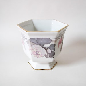 May include: White, octagonal ceramic planter with a gold rim and base. The planter features a floral design with pink and grey flowers and leaves. A decorative piece for indoor plants or home decor.