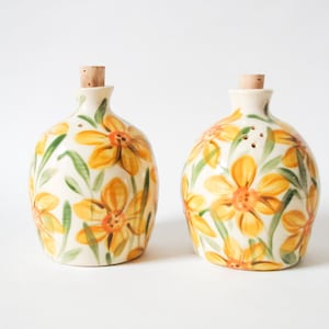 May include: Two ceramic bottles with cork stoppers. The bottles are decorated with yellow and orange flowers and green leaves on a cream background. They are likely for oil and vinegar.