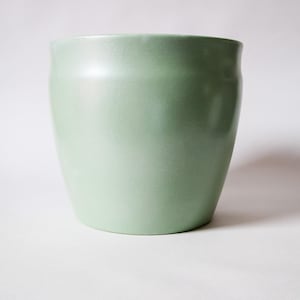 May include: A pale green ceramic plant pot with a smooth, glossy finish. The cylindrical pot has a slightly flared opening and a rounded base. The pot is empty and sits on a white surface.