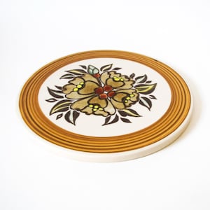 May include: A round ceramic trivet featuring a central floral motif with brown, yellow, and dark green petals and leaves. The design is surrounded by a series of concentric golden-brown rings on a white background, creating a circular border.