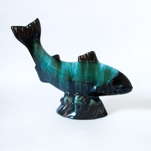 May include: A ceramic fish figurine with a glossy finish. The fish is primarily teal and green with dark brown accents on the fins and tail. It is mounted on a dark brown base, creating a decorative accent piece.