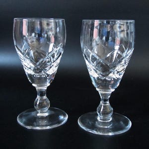 May include: Two clear crystal wine glasses with diamond-cut patterns. The glasses have a rounded bowl, a stem with a bulbous detail, and a circular base. The background is dark.