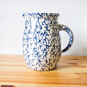 May include: A cream-coloured ceramic jug with a blue splatter pattern. The jug has a large handle and a spout. It is displayed on a wooden surface.