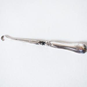 May include: A silver-coloured antique surgical instrument. It features a curved hook at one end and a handle with an engraved design. The instrument is set against a white background.