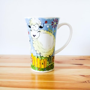 May include: A tall white ceramic mug featuring a cartoon illustration of a fluffy white sheep standing in a green field with a yellow fence. The background is a light blue sky with colorful flowers and the word baaa written in small yellow letters.
