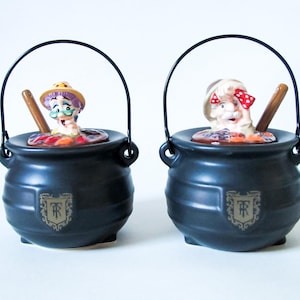 May include: Two black ceramic cauldron-shaped containers with arched handles. Each cauldron features a cartoonish figure with a wooden spoon inside. One figure wears a hat, the other a bow. The cauldrons have a decorative emblem.