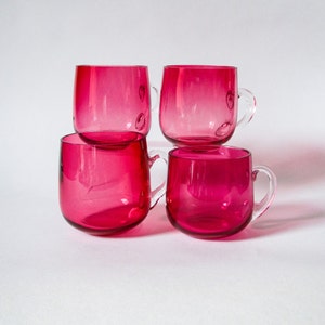 May include: Four clear glass mugs with a pink hue and rounded handles. The mugs are stacked on top of each other.