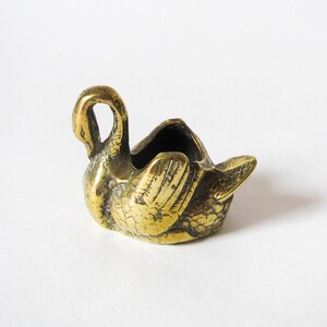 May include: A small, antique brass swan figurine with a hollowed-out body. The swan has detailed feather carvings and a curved neck, with a small opening in the back. The brass has a tarnished, aged appearance.
