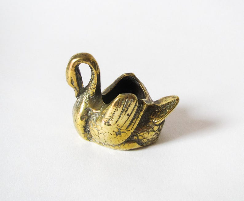 May include: A small, antique brass swan figurine with a hollowed-out body. The swan has detailed feather carvings and a curved neck, with a small opening in the back. The brass has a tarnished, aged appearance.