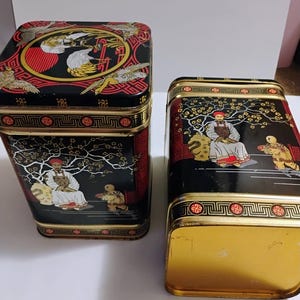 May include: Two rectangular metal storage tins featuring black backgrounds with gold and red accents. The tins are decorated with illustrations of figures in traditional attire and crane motifs. The lids and sides have intricate geometric borders.