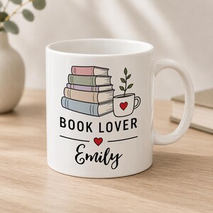 May include: A white ceramic mug featuring a stack of four pastel-coloured books next to a small potted plant. Below the illustration, the text reads BOOK LOVER with a small red heart and the name Emily written in a black cursive script font.