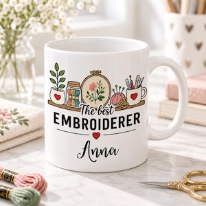 May include: A white ceramic mug featuring a colourful illustration of embroidery supplies including a hoop, thread, and needles. The text reads The best EMBROIDERER followed by the name Anna in black script, with a small red heart icon below.