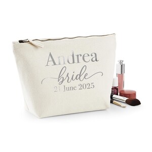 May include: A cream-colored cosmetic bag with the name "Andrea" and the word "bride" printed in silver. The date "21 June 2025" is also printed on the bag. Makeup items, including a brush and lip gloss, are next to the bag.