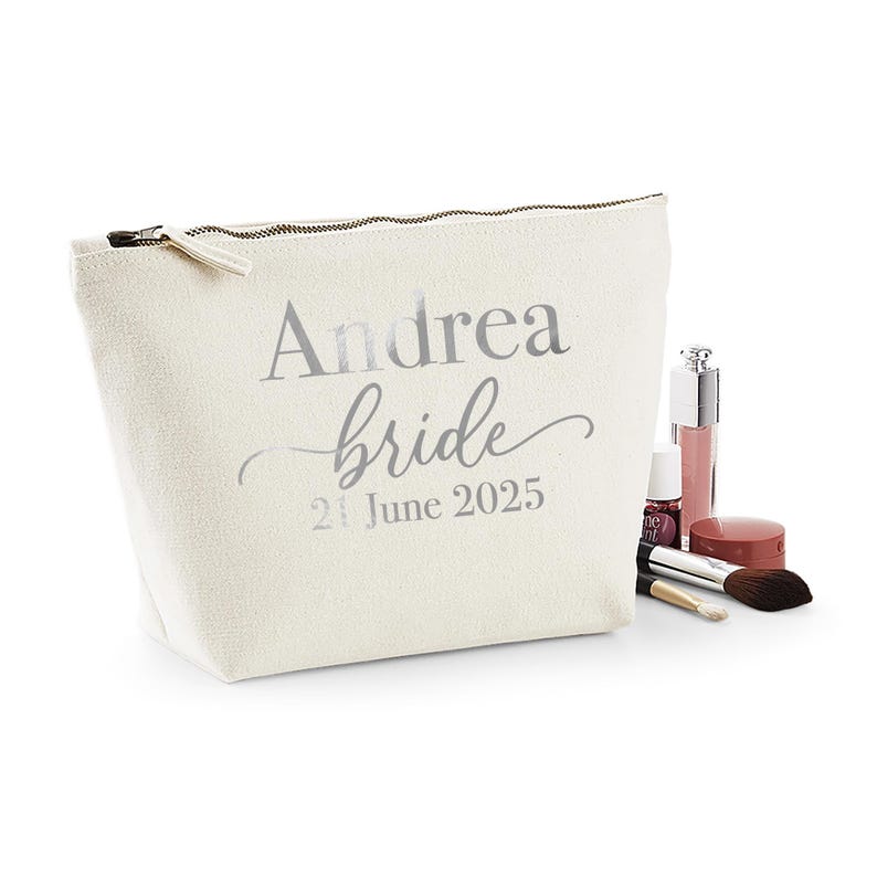 May include: A cream-colored cosmetic bag with the name "Andrea" and the word "bride" printed in silver. The date "21 June 2025" is also printed on the bag. Makeup items, including a brush and lip gloss, are next to the bag.