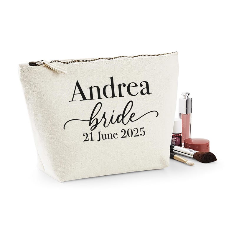 May include: A cream-colored cosmetic bag with a zipper closure. The bag is printed with the name "Andrea", the word "bride", and the date "21 June 2025" in black. Makeup items, including lip gloss and brushes, are next to the bag.