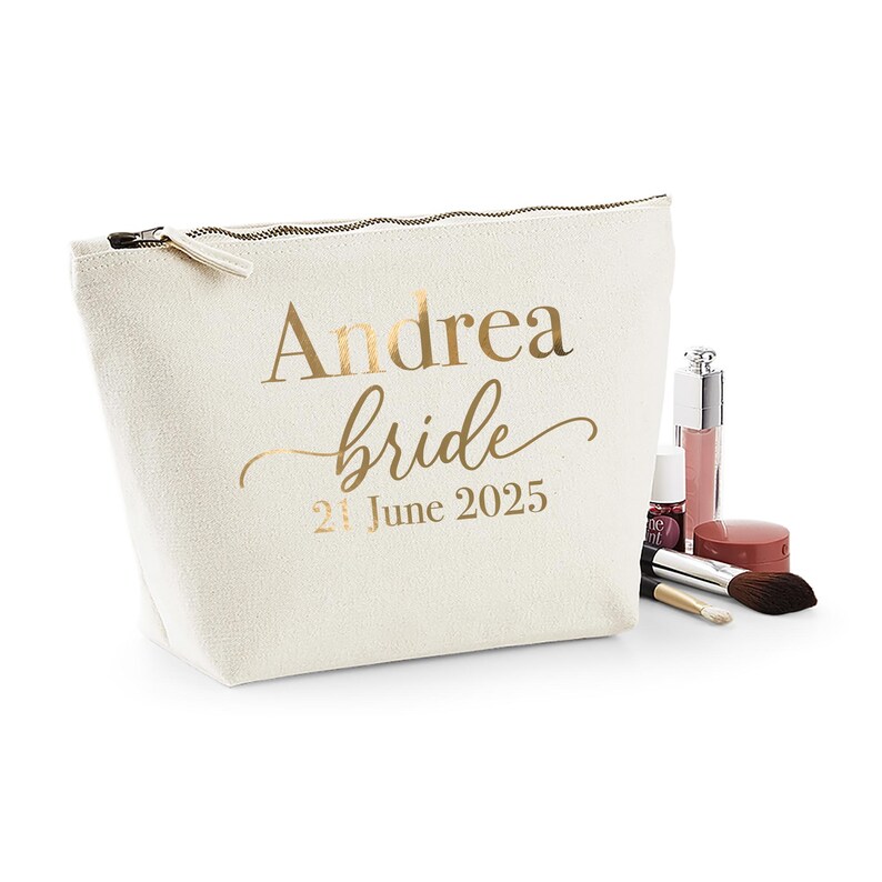 May include: A cream-colored cosmetic bag with a gold zipper and the words "Andrea bride 21 June 2025" printed in gold. Makeup items, including a brush, lip gloss, and eyeshadow, are arranged next to the bag.