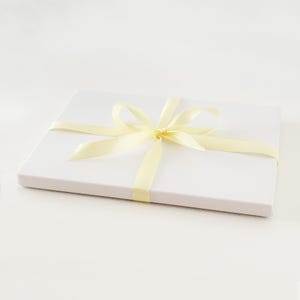 May include: A white gift box tied with a pale yellow satin ribbon. The ribbon is wrapped around the box and tied in a bow on top. The box is rectangular and sits on a white surface.