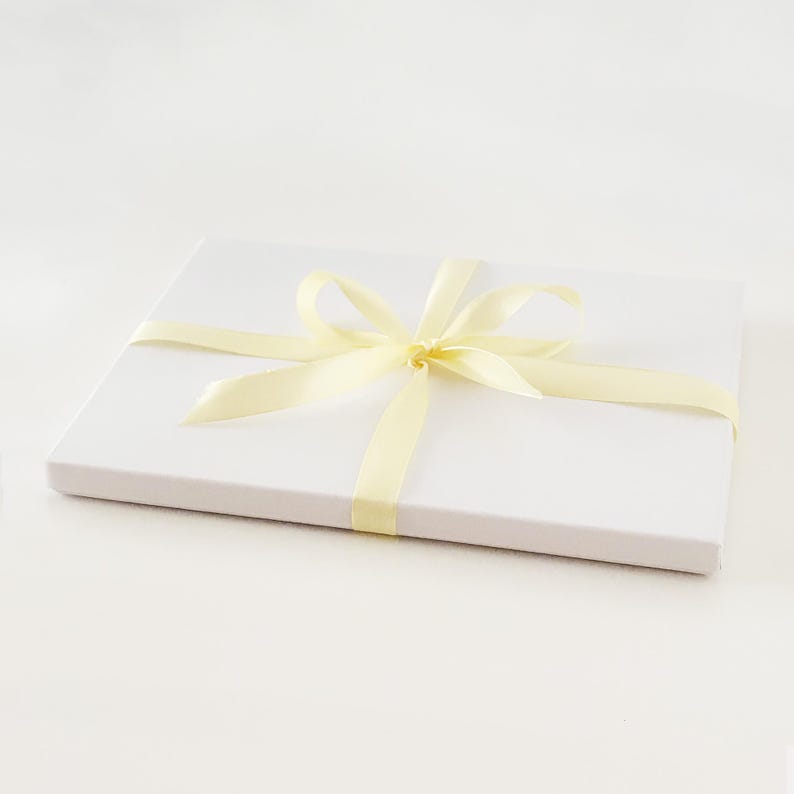 May include: A white gift box tied with a pale yellow satin ribbon. The ribbon is wrapped around the box and tied in a bow on top. The box is rectangular and sits on a white surface.