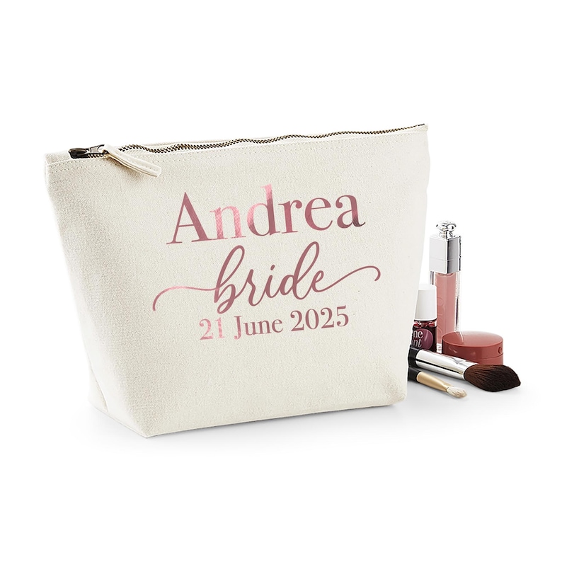 May include: A cream-colored cosmetic bag with a zipper closure. The bag is printed with the name "Andrea", the word "bride", and the date "21 June 2025" in rose gold lettering. Makeup items are arranged next to the bag.