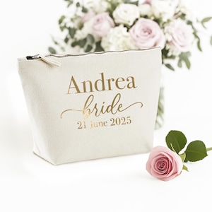 May include: Off-white canvas cosmetic bag with a gold zipper and the text "Andrea bride 21 June 2025". A pink rose and a bouquet of pink and white flowers are in the background. The bag is a wedding-themed item.