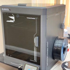 May include: A grey ELEGOO Centauri Carbon 3D printer with a clear door and a small touchscreen display. A spool of black filament is attached to the side. The printer is on a black surface.