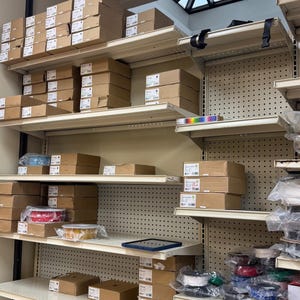 May include: A retail shelving unit with multiple levels holding various brown cardboard boxes, rolls of clear adhesive tape, and plastic-wrapped items. The shelves are made of beige metal with a perforated back panel and a drop ceiling above.