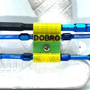 May include: A yellow and green 3D printed holder with the words "DOBRO NEVADA" printed on it. The holder is holding three blue and silver screwdriver bits. The bits are labeled "VICRING H1/4" and "VICRING H7/32".