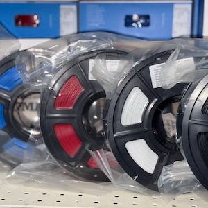 May include: Four spools of filament for 3D printing, wrapped in plastic. The spools are black with different colored filaments: blue, red, white, and black.