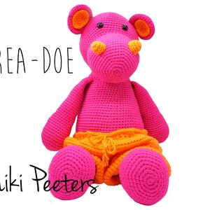 Peut inclure: Un hippopotame en crochet rose portant un short orange. L'hippopotame est assis les jambes croisées. Le texte "KREA-DOE" est écrit en noir au-dessus de l'hippopotame. Le texte "Niki Peeters" est écrit en noir en dessous de l'hippopotame.