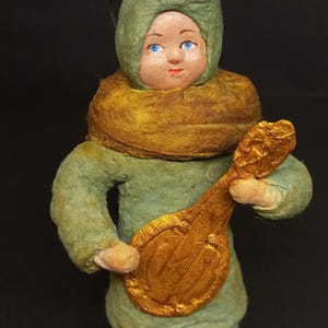 Puede incluir: Figura vintage de una persona con un atuendo verde con capucha a juego y una bufanda dorada. La figura sostiene un laúd dorado. La cara tiene ojos azules y boca roja. Probablemente un adorno navideño.