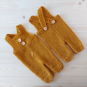 May include: Two mustard yellow knitted baby overalls with wooden buttons. The overalls are made of a soft, chunky yarn and have a simple, classic design.