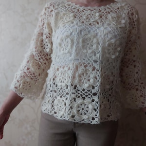 May include: Ivory-colored crochet top with three-quarter sleeves. The top features a floral pattern throughout the design. The crochet work is intricate, creating a delicate and textured appearance. The top is a light, airy garment.