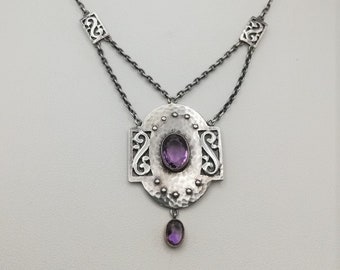Puede incluir: Un collar de plata con un colgante que presenta una amatista ovalada rodeada de un intrincado diseño de plata martillada. El colgante cuelga de una cadena delicada con dos cuadrados de plata más pequeños.