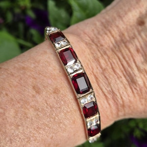 Könnte beinhalten: Ein goldfarbenes Armband mit einer Reihe von rechteckigen, tiefroten facettierten Edelsteinen, die sich mit Paaren kleiner, klarer, runder facettierter Steine abwechseln, welche in einem linearen Muster auf dem Metallband angeordnet sind.