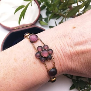 May include: A vintage gold-coloured bracelet with a floral design. It features dark purple, round, glass stones. The bracelet is connected by delicate chains and is displayed on a wrist. The bracelet is in an open, maroon jewellery box.