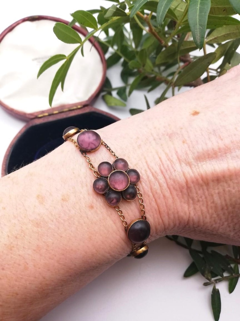May include: A vintage gold-coloured bracelet with a floral design. It features dark purple, round, glass stones. The bracelet is connected by delicate chains and is displayed on a wrist. The bracelet is in an open, maroon jewellery box.