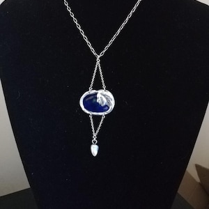 May include: A silver necklace with a blue enamel pendant featuring a stylised face. The pendant is attached to a delicate chain with a small heart charm.