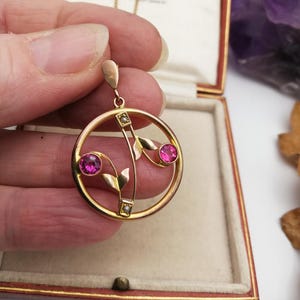 Puede incluir: Un collar con un colgante de oro de diseño circular con dos piedras rosas y una perla. El colgante tiene la forma de un motivo floral estilizado.