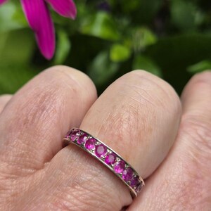 May include: A silver-toned eternity band ring featuring a continuous row of round-cut, vibrant pink gemstones set in a shared-prong metal setting that encircles the entire circumference of the band, creating a uniform and sparkling appearance.