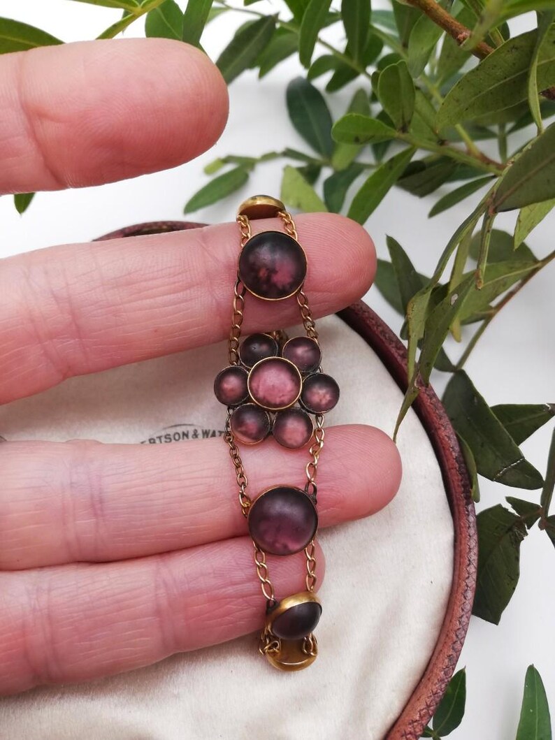 May include: A vintage gold bracelet featuring dark purple and pink cabochon stones. The stones are linked by fine gold chains. The bracelet is held between fingers against a neutral background with some green foliage.