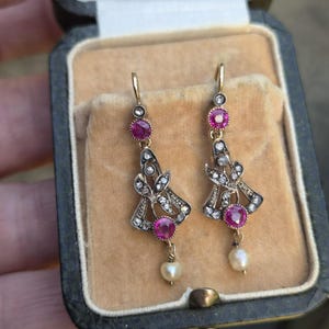 Puede incluir: Un par de pendientes colgantes con piedras preciosas rosas redondas, pequeñas piedras transparentes engastadas en un marco de metal decorativo y una perla blanca que cuelga en la parte inferior de cada pieza, presentados en un joyero abierto.