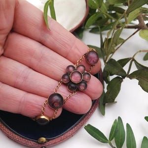 May include: A vintage gold bracelet with a floral design. The bracelet features circular and flower-shaped elements in pink and purple hues. The bracelet is held in a person's hand, with a dark blue box in the background.