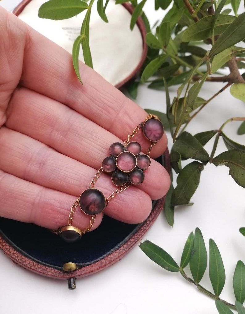 May include: A vintage gold bracelet with a floral design. The bracelet features circular and flower-shaped elements in pink and purple hues. The bracelet is held in a person's hand, with a dark blue box in the background.