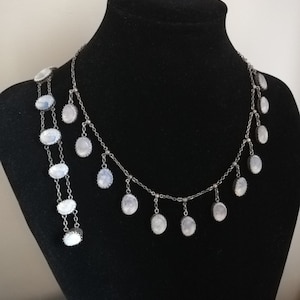 May include: A silver chain necklace with oval-shaped moonstone pendants. The necklace has a delicate design and features multiple strands of chain.