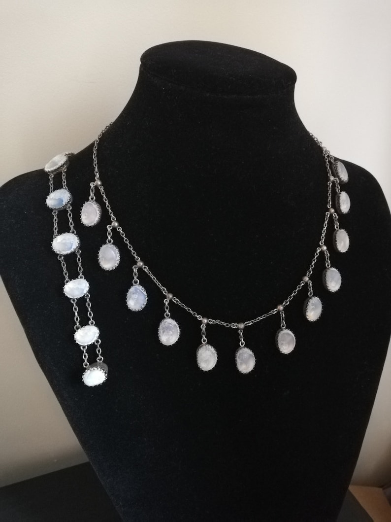 May include: A silver chain necklace with oval-shaped moonstone pendants. The necklace has a delicate design and features multiple strands of chain.