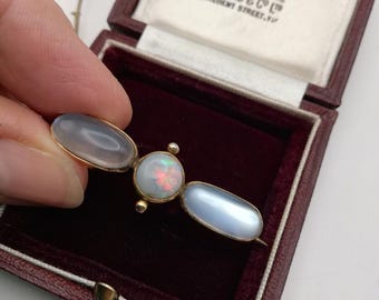 Könnte beinhalten: Vintage goldene Brosche mit drei Edelsteinen: einem ovalen grauen Stein, einem runden Opal und einem hellblauen ovalen Stein. Die Brosche wird in einer bordeauxfarbenen Schatulle mit dem Text "By appointment to H.M. The Queen" präsentiert.